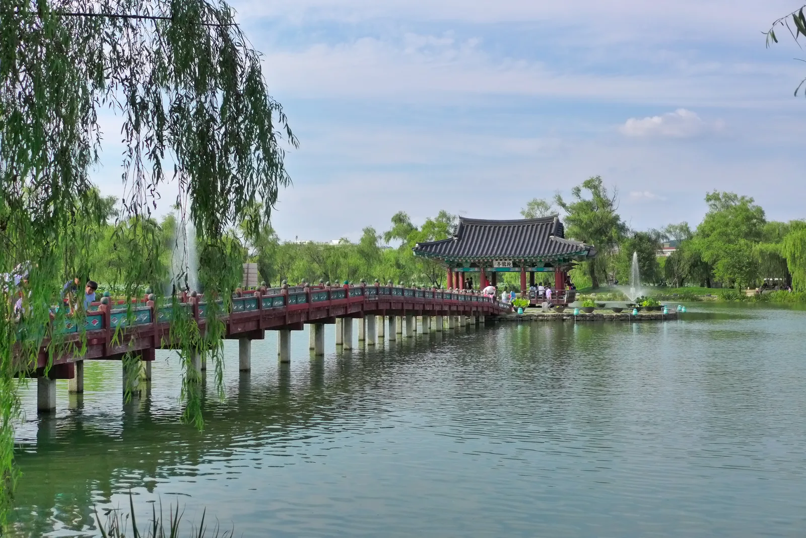 A curved bridge crosses Gungnamji Pond toward Poryongjeong pavilion among willow trees and fountains in Buyeo