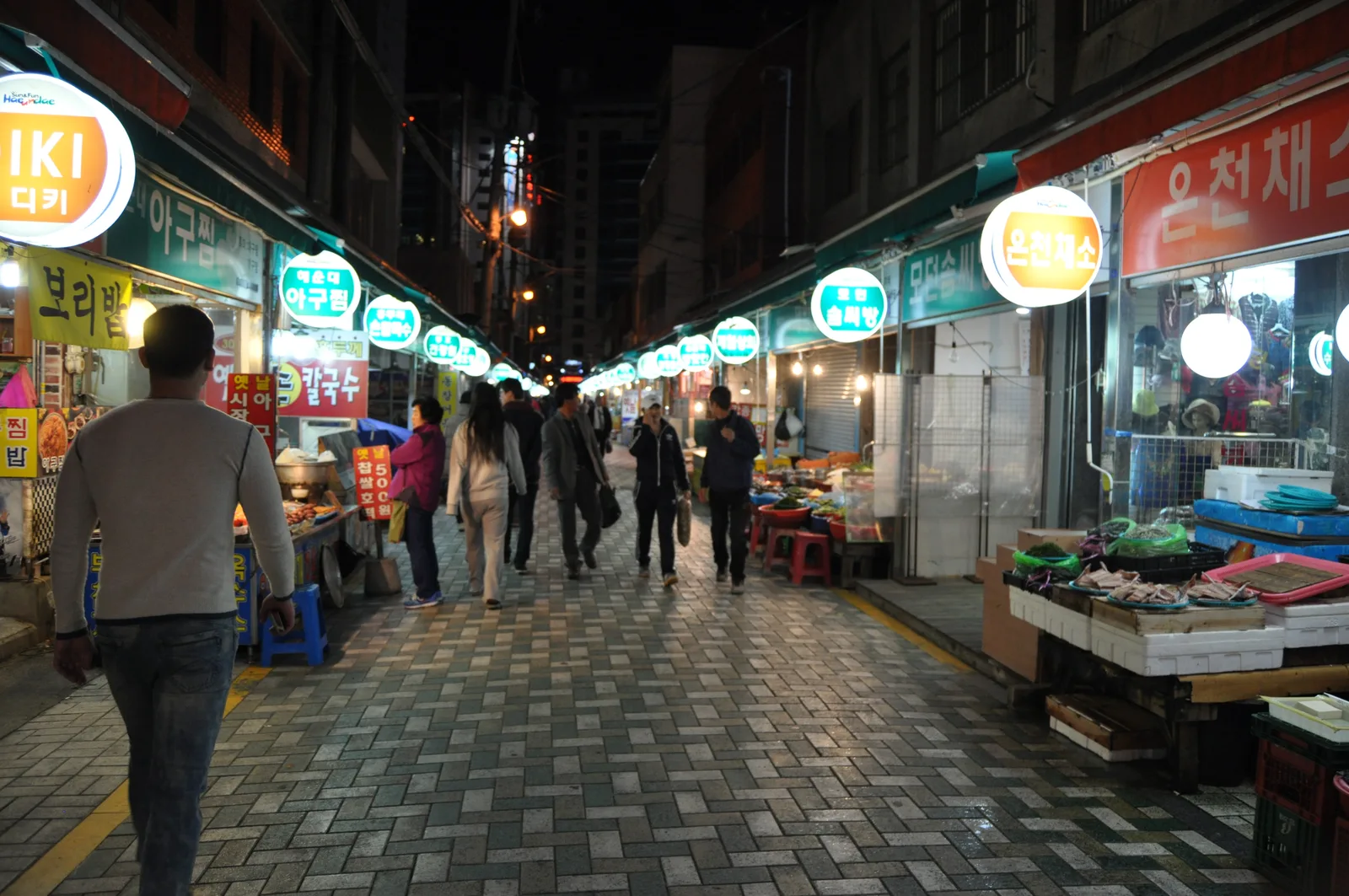A narrow evening lane with illuminated food signs, seafood counters and shoppers inside Haeundae Traditional Market