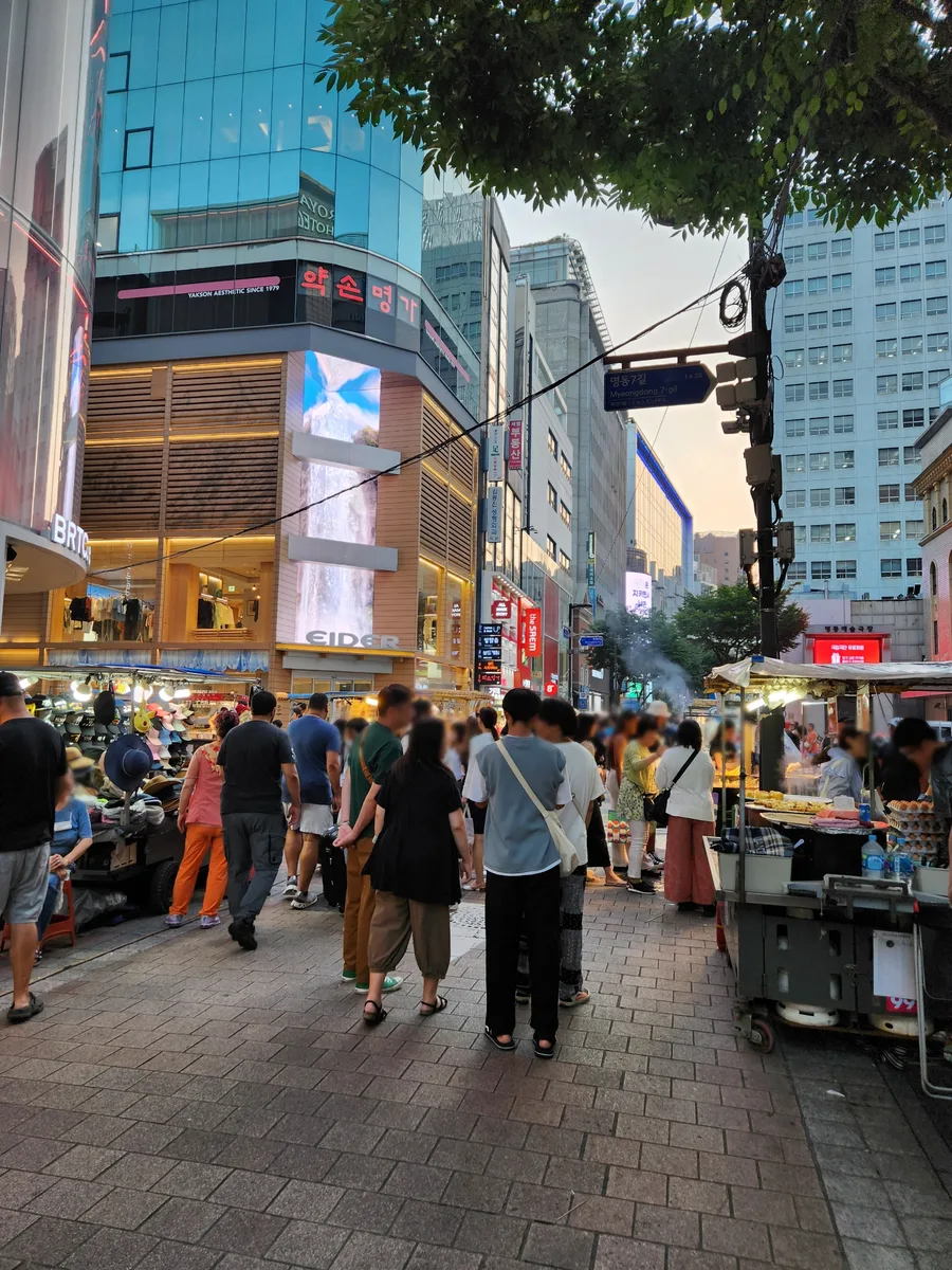 Travelers walking between beauty shops and street-food stalls in Myeongdong