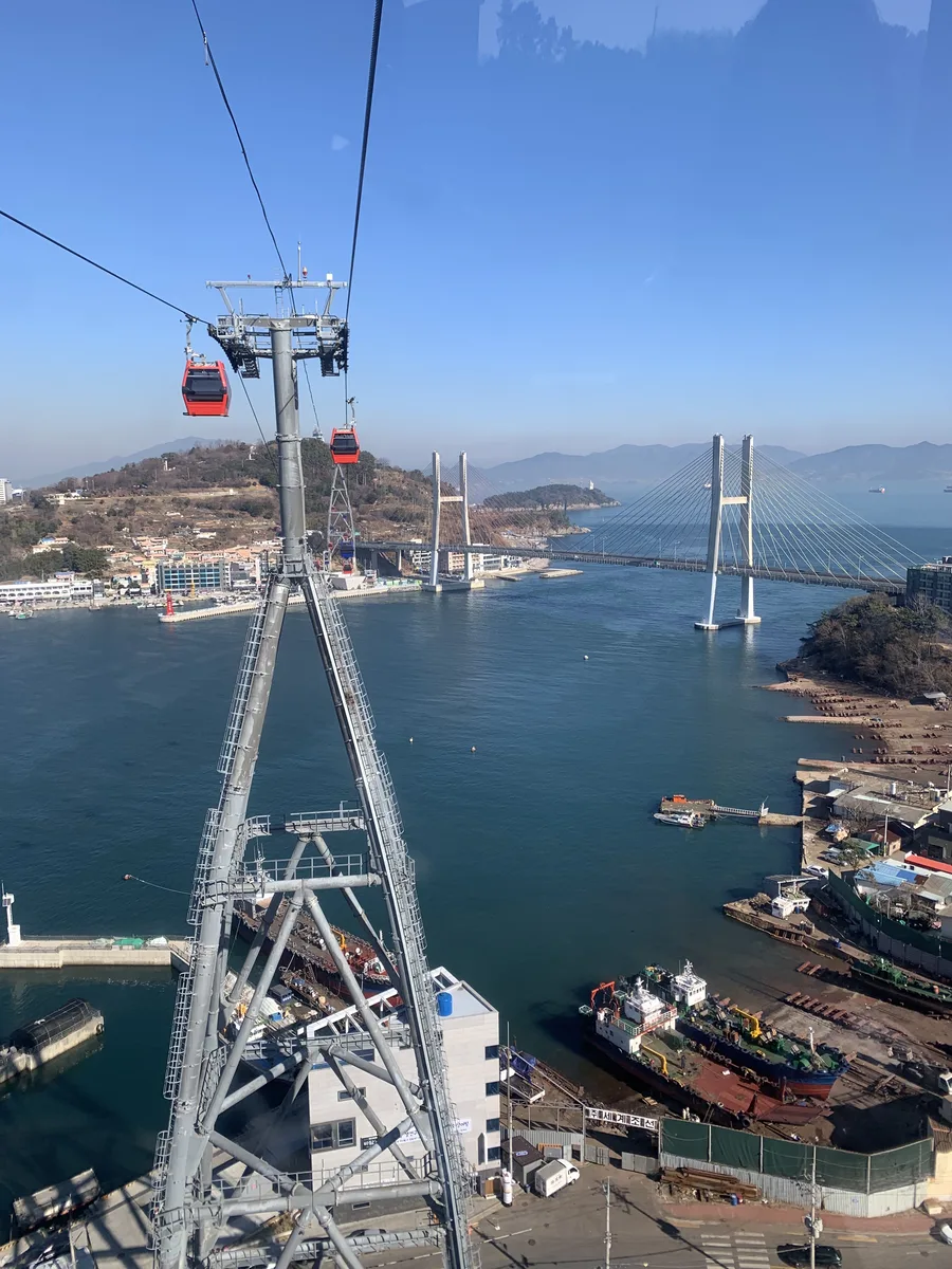 Red Yeosu Maritime Cable Car gondolas crossing above the harbor, terminal roofs and Dolsan Bridge