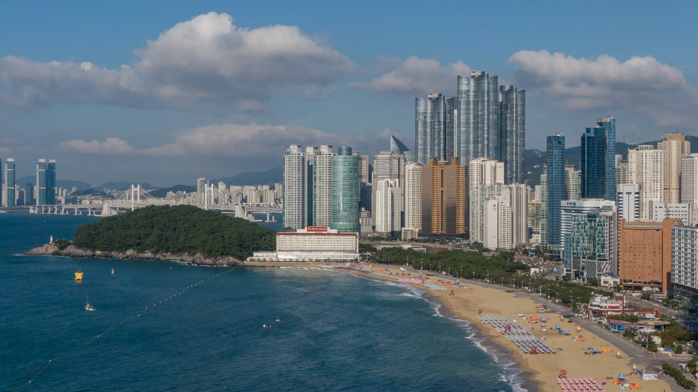 Aerial panorama over the curve of Haeundae Beach, the waterfront skyline, Dongbaekseom and distant Gwangan Bridge
