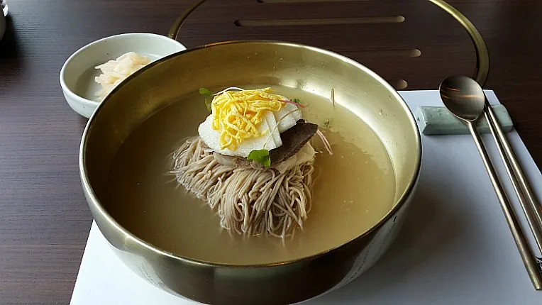 A metal bowl of naengmyeon with cold broth, noodles, sliced meat, egg and cucumber