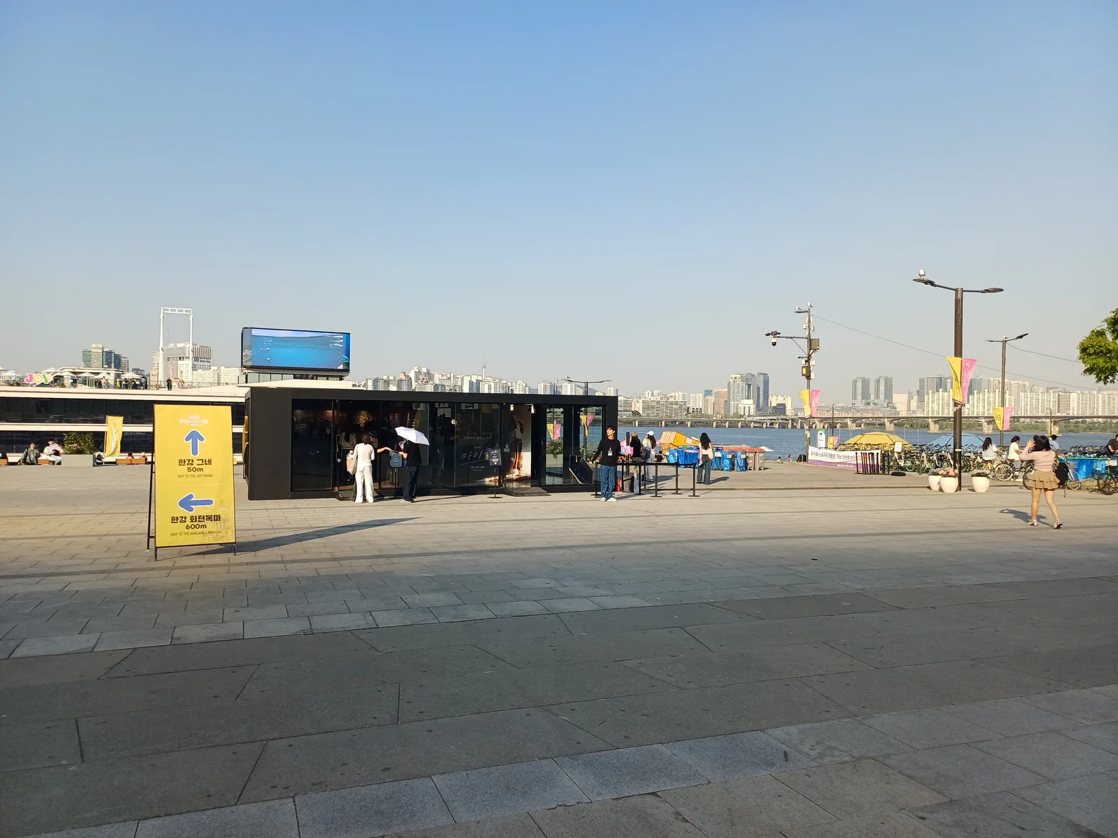 Visitors crossing a broad riverside plaza toward the Han River and Seoul skyline under a clear sky